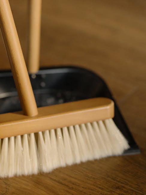 A woman with light brown hair tied back, dressed in a lavender long-sleeve top and yellow cleaning gloves, is performing surface cleaning on a wooden panel in a room with neutral grey walls and ample 